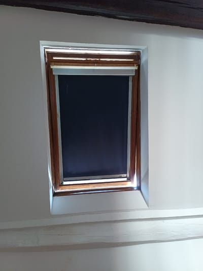 A slightly angled medium shot shows a skylight window with a dark roller blind partially drawn. The window is framed by a wooden casing and set into a white, textured wall. Above the window, a dark wooden beam runs horizontally across the top of the frame. Below the window, two more white, textured beams are visible, one directly beneath the skylight and another lower down. The scene appears to be indoors. The location of the image is identified as Montgeron, France.