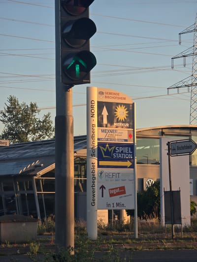 The image captures a street scene in Bischofsheim, Germany, featuring a traffic light with a green arrow illuminated, indicating the direction of travel. A prominent signpost is present, displaying multiple advertisements and directional information. These include signs for "GEMEINDE BISCHOFSHEIM," "MERKUR," "MEXNER GmbH," "SPIEL STATION," and "REFIT." Another sign points towards "Bischofsheim." In the background, a building with a modern facade and large windows is visible, along with power transmission towers and overhead wires, suggesting an industrial or commercial area. Overgrown vegetation and a concrete structure are also present in the foreground, indicating a somewhat untended outdoor space. The overall lighting suggests it is either early morning or late afternoon.