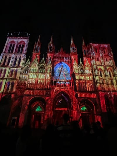 The image depicts the Cathédrale Notre-Dame de Rouen in Rouen, France, at night, serving as the canvas for a light projection show. The grand Gothic facade of the cathedral is illuminated with vibrant, painterly projections. Predominant colors include deep reds, oranges, yellows, and greens, accentuating the intricate architectural details, spires, and arched windows. A large central rose window is distinctly highlighted in a brilliant blue hue, revealing its internal structure. Several lower arched entrances feature luminous green illumination. In the foreground, numerous silhouetted individuals are visible, facing the cathedral and observing the light display. The sky above is entirely black, indicating a nighttime setting.