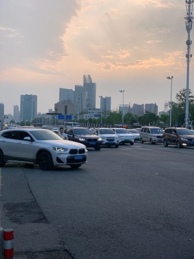 The image shows a street in Wuhan, China (latitude: 30.51, longitude: 114.404) with several vehicles stopped in traffic. There are at least six vehicles in the image, including a white BMW SUV in the foreground. One person is visible, a man standing next to a black BMW SUV. The scene appears to be a street in the city, with buildings in the background. The sky is overcast and the time appears to be late afternoon. The road is paved and appears to be a main thoroughfare with several lanes. The image suggests that the traffic is heavy and may be congested.