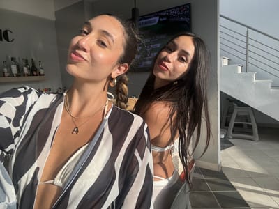 The image shows two young women posing for a selfie. The woman on the left is closer to the camera, smiling with her head tilted. She is wearing a black and white striped sheer cover-up over a white bikini top and a gold necklace with an "A" pendant. The woman on the right is slightly behind and to the right, also looking towards the camera. She has long, dark hair and is wearing a white bikini top. Behind them, a television screen displays a sports broadcast. A shelf with bottles is visible on the left wall. To the right, a modern staircase with a silver railing leads upwards. The setting appears to be an indoor or semi-outdoor area with tiled flooring and natural sunlight casting shadows. The overall atmosphere suggests a casual, possibly vacation or leisure setting. The location is identified as San Juan, United States of America.
