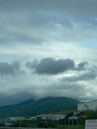 The image depicts an overcast sky with thick, dark clouds dominating the view. The sky occupies most of the frame, with a small section showing a green landscape and some buildings, possibly within the urban area of Gorovani, Georgia. A sign with Georgian text is visible on the right side of the frame. The image is taken at an angle, not perfectly horizontal. There are no discernible people or specific activities visible.