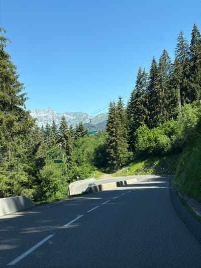 The image depicts a winding asphalt road in Châtel, France, a mountain village. The road, marked with white dashed lines, curves to the right and ascends a gentle slope. On the left side of the road, a concrete barrier lines the edge, with lush green trees and tall conifer trees growing beyond. In the background, a ski lift with green seats is visible, suspended by cables stretching across the frame, suggesting a mountainous terrain common in ski resorts. Beyond the trees, snow-capped mountains rise against a clear blue sky. The environment is characterized by dense forestation and a bright, sunny day.
