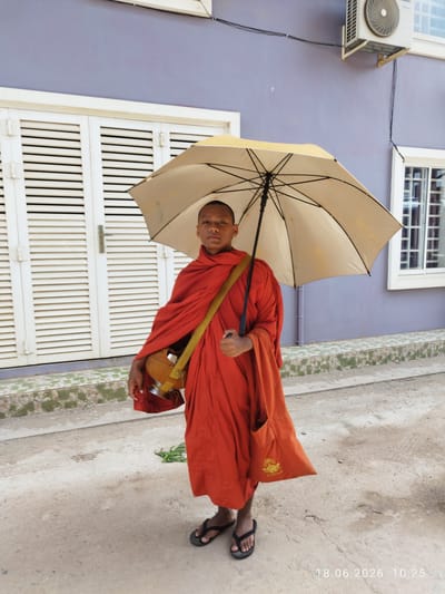 A young Buddhist monk in orange robes stands on a dusty road in front of a building with a lavender-colored wall and white louvered doors. He holds a light-colored umbrella over his head. A yellow strap crosses his chest, supporting a round alms bowl. An orange tote bag hangs from his right shoulder. The monk is wearing black flip-flops. To the right of the monk, a window with white mullions is visible on the building. In the lower right corner of the image, a date and time stamp reads "18.06.2026 10:25". A green sprig of vegetation lies on the ground near the monk's feet. The building appears to be located in Kampot, Cambodia, based on the provided context.