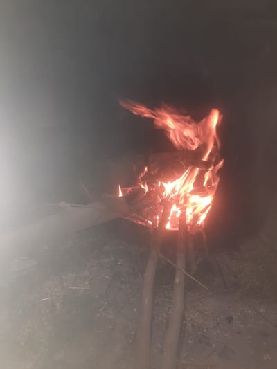 A bonfire burns in a dark, outdoor environment. The fire is contained within a makeshift structure of logs, with flames leaping upwards and casting an orange glow. Smoke billows around the fire, particularly on the left side of the image, creating a hazy effect. The ground beneath appears to be composed of dirt and debris. The scene is dimly lit, suggesting it is nighttime. No people are visible in the image. The location is identified as Voseaabad, Tajikistan.