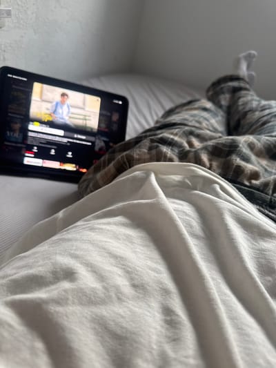 A person lies in bed, wearing a plaid shirt and white t-shirt. Their legs are bent, feet visible in white socks. Next to them is a tablet displaying a streaming service interface with a person on the screen. The setting appears to be a bedroom, with white bedding adding to the relaxed atmosphere.