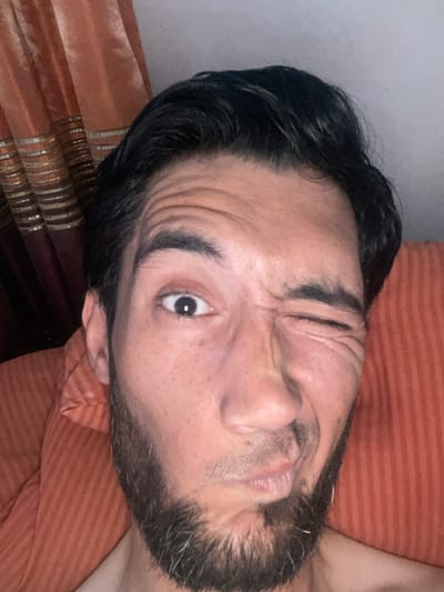 The image is a close-up, selfie-style photograph of a man's face, with his left eye open and his right eye squinting or closed. He has dark hair and a beard. He is positioned in front of an orange, textured background, which appears to be upholstery or a curtain. The man's facial expression is playful or humorous, with his mouth slightly open and a wrinkle on his forehead. The lighting is somewhat dim, creating shadows on his face. The image does not contain any visual information that would indicate its location in Mashrab, Uzbekistan.