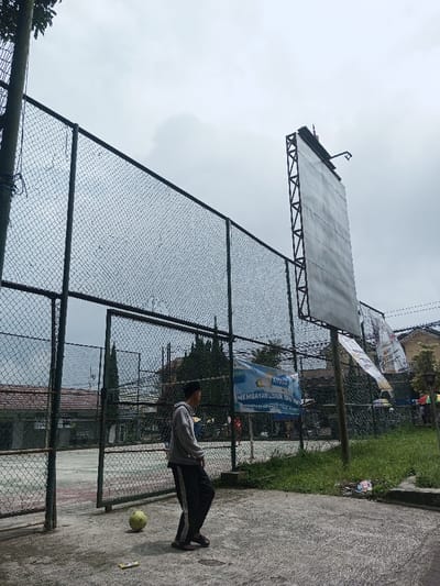 An overcast day reveals a recreational area with a chain-link fence surrounding a concrete surface. A young man stands on the concrete near a yellow soccer ball. Behind the fence are buildings and various banners, including one that says "MEMBAYAR LISTRIK TEPAT WAKTU!". To the right, a tall, rectangular structure stands near a grassy area. The sky is gray and cloudy.