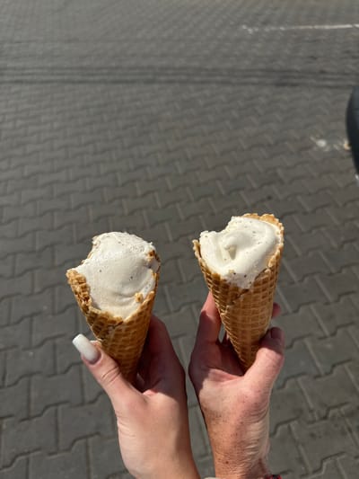 In Mamaivtsi, Ukraine, two hands hold two ice cream cones with bitten tops, the ice cream appearing to be vanilla flavored with visible specks. The hands are against a backdrop of gray, interlocking paving stones under a bright, sunny sky. The person on the left has long, square, white-painted fingernails. The person on the right has freckled skin and a colorful bracelet on their wrist.