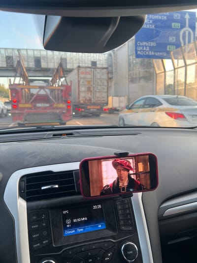 The image shows the interior of a car looking out through the windshield. The car is stopped in traffic, indicated by the presence of other vehicles, including trucks with containers and a white sedan. A road sign with Russian text and directional arrows is visible in the background, suggesting the location is near the city of Selyatino, Russia, as indicated by the text "СЫРЬЕВО" and "МОСКВА-ЦЕНТР". On the dashboard, a smartphone is mounted and displaying a video of a man in historical attire. The car's radio display shows the time as 20:26 and a radio station frequency of 90.50 MHz. The overall environment is an urban highway or road.