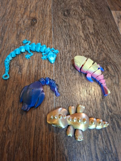 The image shows four 3D-printed articulated toys on a wooden surface. From left to right, the toys are a turquoise lizard, a multi-colored fish, a dark blue and purple bird, and a metallic gold scorpion. The wooden surface appears to be a table or floor with a wood grain pattern. There are no people or signs of human activity in the image. The image does not provide any information about the location in the city of Gorkovskiy, Russia.