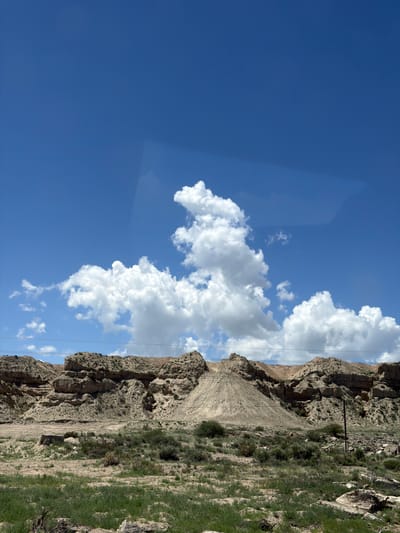 The image shows a vast, arid landscape under a bright blue sky with scattered white clouds. In the foreground, dry, sparse vegetation grows among rocks and sandy soil. A pile of light-colored dirt or gravel is visible on the left side of the frame, with a steep, rocky incline forming the background. Power lines are strung across the scene, supported by utility poles, indicating human infrastructure in this remote area. The terrain is characterized by rugged rock formations and erosion, typical of a desert or semi-arid environment. There are no people visible in the image. The location is identified as Tong, Kyrgyzstan.