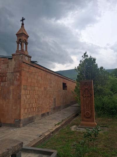 A stone church with a bell tower topped with a cross stands under a cloudy sky in Martiros, Armenia. The church is constructed of red-orange stone bricks and has a narrow pathway leading away from it. To the right of the pathway is a tall, intricately carved stone monument with a cross and ornate designs. Green trees and lush vegetation are visible behind the monument and to the right of the church, with rolling hills in the distance. The scene appears to be an outdoor religious site.