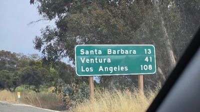 A green highway sign indicates distances: Santa Barbara 13, Ventura 41, Los Angeles 108. It's mounted on wooden posts beside a road, with dry grass and foliage around it. A tree looms behind. The sign is partially obstructed by a dark object on the right side.