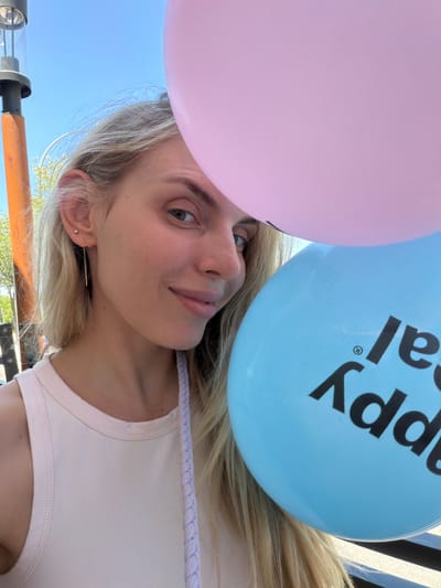 A young woman with blonde hair and blue eyes is partially visible in the frame, holding two balloons. She is wearing a light pink tank top and a delicate gold earring. Her face is turned to the side, and she has a slight smile.
One balloon is pink, and the other is light blue with black lettering. The blue balloon has the partial text "appy" and "al" visible, suggesting it says "Happy Birthday". The image appears to be taken outdoors on a sunny day, as indicated by the bright lighting and the clear blue sky in the background. There are some trees and structures in the background, which are blurred. The overall mood is cheerful and celebratory. The woman's hair is flowing, and she appears to be looking up. The scene is shot from a low angle, with the balloons dominating the upper portion of the image.