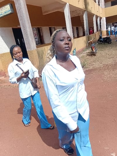 Two women stand on reddish-brown ground near a building. Both are wearing white long-sleeved shirts and blue pants. The woman closer to the camera holds a mobile phone. The other one appears to be looking upward. In the background, a building is visible with multiple windows and balconies. There's a motorbike parked in front of the building. The sky is clear and the lighting is bright, suggesting it's daytime.