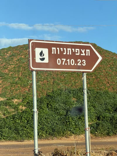 A brown arrow-shaped sign with white Hebrew text "תצפיתנין" and "07.10.23" is mounted on two metal poles. A white symbol of a flame within a capsule is at the bottom. Lush green vegetation covers a hillside behind the sign. A dirt path runs parallel to the right pole. A clear blue sky with thin white clouds is visible in the upper left corner.
