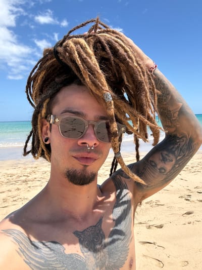 A sun-drenched portrait captured on a beach in Pájara, Spain, features an individual with dreadlocks, sunglasses, and extensive tattoos. The person is photographed from the chest up, seemingly reclining or lying on their back, looking directly towards the camera.
The subject has long, multi-toned brown dreadlocks, some adorned with small silver-toned beads or wraps. They wear stylish, grey-tinted sunglasses that reflect the bright blue sky and sandy beach. A prominent silver septum piercing and two small silver nostril studs are visible, complementing a short goatee. A dark earring is seen in the left ear, and dark nail polish is applied to the visible fingernails. A small cross symbol is visible on the arm of the sunglasses.
Intricate black and grey tattoos cover both arms and a portion of the chest. The left arm features a detailed portrait, possibly of a woman's face, alongside other complex designs. The chest and right shoulder display a large tattoo of a bird, possibly a raven or similar, with prominent wings. A red and white braided bracelet is worn on the wrist of the raised left arm.
The setting is a vibrant beach under a clear, bright blue sky with scattered white clouds. A calm, turquoise ocean with gentle waves and white foam lines is visible in the middle ground, while light brown sand with numerous footprints fills the foreground. The strong daytime sunlight creates crisp shadows and highlights, enhancing the textures of the dreadlocks and the details of the tattoos. The overall impression is one of relaxation and enjoyment in a beautiful, sunny coastal environment.