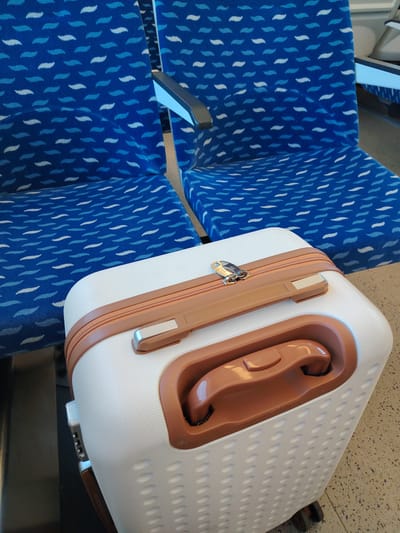 A close-up shot shows a white suitcase with brown accents placed on the floor in front of two empty blue seats on a train. The seats are upholstered in a fabric with a wave-like pattern in varying shades of blue and white. The suitcase is a hard-shell model with a textured surface and a recessed handle. A brown zipper runs along the top edge of the suitcase, secured by a silver lock with the "YKK" logo visible. The scene appears to be set inside a train carriage, likely during daylight hours given the ambient lighting. There are no people visible in the frame, suggesting the train is either stopped or passengers are not in the immediate vicinity of the camera. The overall impression is one of travel and anticipation, with the empty seats implying a journey about to begin or conclude. The location context of Ulm, Germany, suggests this could be a regional or long-distance train within Germany. The image is well-lit, with no discernible weather conditions.