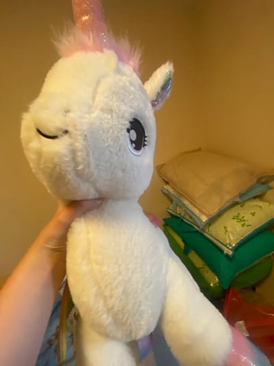A person's hand is holding a white plush unicorn toy. The unicorn has a pink, glittery horn and pink accents on its hooves and ears. The unicorn's eyes are closed, giving it a peaceful expression. In the background, a stack of pillows is visible, some of which are green and yellow. A red plastic bag is also present in the background. The scene appears to be indoors, possibly in a bedroom or storage area. The image does not provide any information about the city of Afanasyevo, Russia.