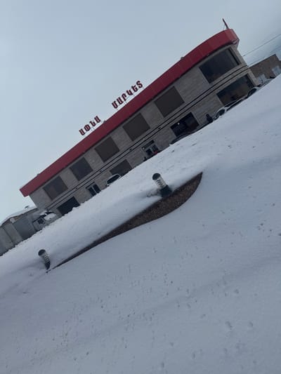 The scene depicts a snow-covered urban environment in Apnagyugh, Armenia, under an overcast sky. In the foreground, undisturbed white snow blankets the ground, exhibiting various small depressions and faint tracks. A low, curved divider constructed from light brown aggregate material cuts across the snow, partially obscuring two dark-colored, cylindrical bollard lights with clear, ribbed tops.
The midground features a two-story commercial building with a light gray, textured facade. A prominent red band forms the roofline and extends horizontally along the upper section, displaying the Armenian text "ՍԱԹԵՆ" in red letters. The building contains multiple large, rectangular windows, most of which have dark coverings, and at least two ground-level entrances.
Two individuals are visible near the building's rightmost entrance. One person, dressed in dark clothing, stands near a parked, light-colored sedan that is partially covered by snow. Another individual is partially obscured near the entrance. A second light-colored vehicle, possibly a van or truck, is parked further to the left of the building. The background shows additional snowy structures and a featureless, light gray sky.
