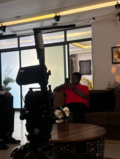 In Pakuro, Nigeria, a young Black man in a red shirt and black pants is seated on a brown sofa, engrossed in his smartphone. The scene is indoors, with a camera on a tripod in the foreground, suggesting a film or video recording is in progress. The room features large windows, sleek ceiling lights, and a round wooden coffee table with a vase of flowers. Other individuals are partially visible, one in black pants and shoes to the left, and another, also in black, partially obscured to the far left. The atmosphere is dimly lit, with the natural light from the windows creating a soft glow.