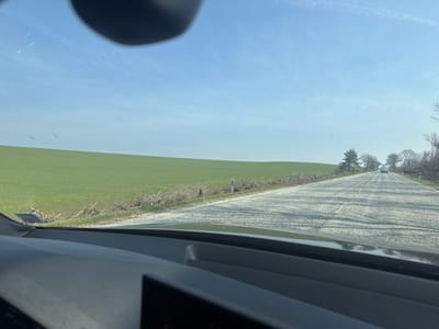A frontal view from inside a moving vehicle depicts a rural road scene in Kozar Belene, Bulgaria, under a clear blue sky with faint contrails. The vehicle's dark dashboard and a portion of a digital display are visible in the lower foreground. A dark, possibly circular object partially obscures the sky in the upper center of the frame. To the left, a vast, gently undulating green field, likely agricultural, extends to the horizon. The field is bordered by a narrow dirt shoulder strewn with dried woody debris, and a white and black roadside marker post is present on this side. The asphalt road, showing visible wear and light-colored patches, features a faded double solid white centerline and recedes into the distance. A white sedan-like vehicle is visible ahead on the road, traveling in the same direction. To the right, the road is flanked by a sparse line of bare deciduous trees and one prominent evergreen tree. No individuals are visible.