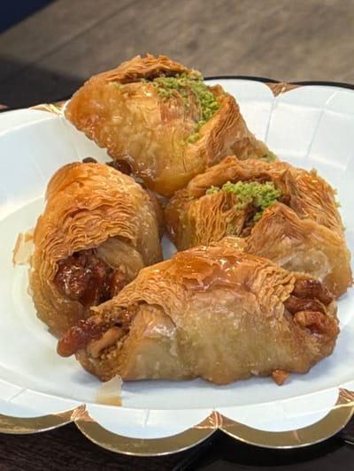 The image displays a close-up, eye-level view of four pieces of baklava-style pastry arranged on a disposable white paper plate with a scalloped edge and a metallic gold border. Each pastry exhibits a golden-brown, flaky, layered phyllo dough exterior, visibly coated with a reflective, clear syrup. The top-right and center-right pastries are topped with a scattering of finely crushed green pistachios. The center-left and bottom-center pastries reveal a dark, chunky nut filling, consistent with walnuts or pecans, partially exposed from within the pastry folds. The plate is positioned on a dark, out-of-focus surface, likely a table or countertop. No individuals or specific actions are depicted. The image does not contain any discernible environmental features or landmarks to determine its specific location within Connah's Quay, United Kingdom, or any other geographical area.