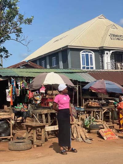 In Ipota, Nigeria, a woman wearing a pink shirt and black skirt stands at a roadside stall. The stall is covered by a striped umbrella and is stocked with various goods including snacks, packaged foods, and personal care items. Behind her, there are piles of yams and coconuts. The stall is constructed with wooden materials and incorporates old tires as part of its structure. In the background, there is a building identified as an Assemblies of God Church. The sky is clear and blue.