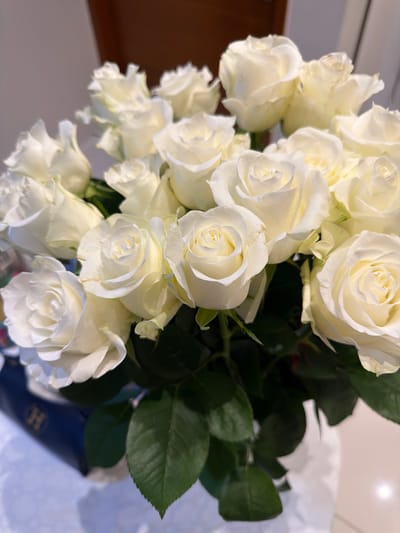 A close-up shot features a bouquet of white roses, some fully bloomed and others still in bud. The roses are arranged tightly, with lush green leaves peeking through. The lighting suggests it's daytime, with a soft glow illuminating the delicate petals. The background is slightly blurred, hinting at an indoor setting, possibly a table or a surface, with a hint of a brown wooden element to the left. The overall mood is serene and elegant, highlighting the natural beauty of the flowers. There is no visible text in the image.