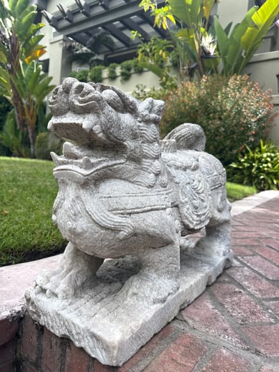 A close-up, eye-level, medium shot captures a weathered, light gray, stone statue of a mythical creature, possibly a foo dog or guardian lion, positioned outdoors on a brick pathway. The statue is predominantly in the foreground, facing left, with its mouth open and sharp teeth visible. Its body is covered in carved decorative patterns and fur textures. The statue rests on a rough-hewn stone base, which sits on a red brick path. Behind the statue, a vibrant green lawn is visible to the left, and lush green foliage with some red-flowered bushes occupies the mid-ground and background. Further back, a modern building with an overhang supported by dark beams is partially visible, along with more greenery on balconies and roofs. The scene suggests a well-maintained outdoor space, likely a residential or commercial property, in Piedmont, California, United States of America. There are no people or animals present in the image, and the lighting suggests it was taken during daylight.