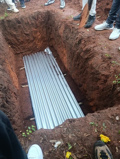 The scene depicts an open burial site in the city of Muthetheni ward, Kenya. A rectangular grave, approximately 2.5 meters deep, has been excavated into reddish-brown soil, revealing rough internal walls. The excavated earth forms mounds around the perimeter of the grave. Laid horizontally across the floor of the grave are multiple sheets of corrugated galvanized iron, which rest on several horizontal wooden beams embedded into the grave walls. The lower legs and feet of at least five individuals are visible, standing on the earthen ground surrounding the grave. Footwear includes white sneakers, black dress shoes, and multi-colored athletic shoes. One individual's hand, located near the top right of the frame, holds the wooden handle of a metal shovel, with its blade angled towards the ground adjacent to the grave's edge. The surrounding ground features sparse green vegetation, dry leaves, and scattered white and yellow floral petals.