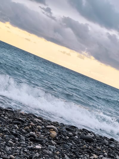 The image captures a slightly tilted, close-up view of a pebble beach on the coast of Zemo Kvariati, Georgia. A wave is breaking onto the shore, with white foam and spray. The water is a deep blue-green, with ripples and texture. Above the water, the sky transitions from a pale yellow near the horizon to dark gray clouds. The beach is covered in a dense layer of smooth, dark-colored pebbles of varying sizes. There are no people or man-made structures visible in the frame. The overall impression is one of natural coastal scenery under an overcast sky.