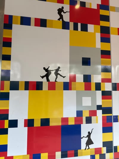 A high-angle shot depicts a section of a large, flat surface adorned with a design reminiscent of Piet Mondrian's abstract style, featuring rectangular blocks of red, yellow, blue, white, gray, and black. The design is not a perfect replication of Mondrian's work, as it includes narrower stripes of color framing larger rectangular areas.
Superimposed onto this geometric background are several black silhouettes of people in acrobatic poses or in the act of falling. One silhouette appears to be dangling by an arm. The image's location is in Best, Netherlands.