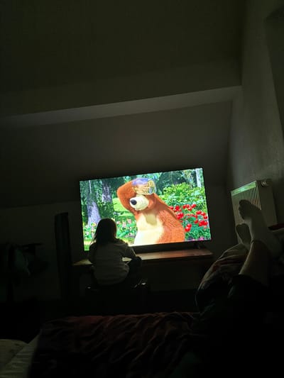 The scene depicts a dimly lit interior room in Herxheim, Germany. A large, flat-screen television dominates the center, displaying an animated program featuring a brown bear character with a clear glass inverted over its head, set against a vibrant outdoor background of green trees and red berry bushes. In the foreground, a young individual, likely a child, is seated on a low surface with their back to the viewer, actively observing the television. The child has long, dark hair and is wearing a light-colored top. To the right, the lower legs and feet of another individual, clad in light-colored socks, are visible, indicating a reclining posture on a bed or sofa in the immediate foreground. A white, panel-style radiator is mounted on the wall to the right of the television. The room's primary illumination originates from the television screen, casting a glow on the child and immediate surroundings, while the remainder of the space, including an angled ceiling on the right, remains in deep shadow.