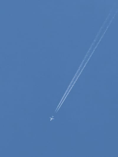 A small white airplane, leaving a prominent contrail, is seen flying diagonally across a clear, bright blue sky. The contrail consists of two distinct, parallel white lines that stretch from near the top right of the frame downwards. The airplane is positioned in the lower middle section of the image. The sky is uniformly blue, indicating daylight with no clouds visible. The image was taken from the ground looking upwards.