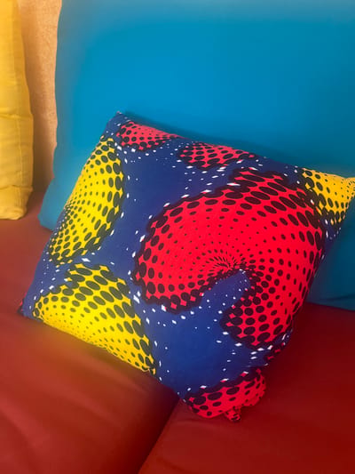 A square throw pillow is displayed with a vibrant, patterned fabric cover. Red, blue, and yellow sections are adorned with black polka dots that give a sense of depth. It rests partially on a teal cushion, with the remaining section on a red surface. A corner of a yellow pillow is also visible beneath.