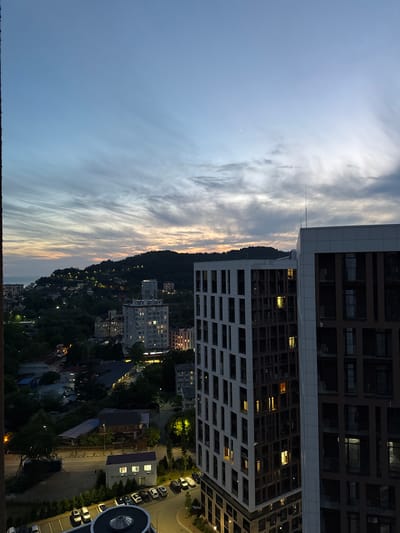 The image is a high-angle, medium shot of a city landscape in Vardane Microdistrict, Russia, during dusk. The sky is a gradient of pale blue and muted orange, with wispy clouds stretched across it, indicating the end of the day. In the foreground, two modern high-rise buildings dominate the right side of the frame. The building on the left has a white facade with a grid of dark windows, some of which are illuminated with a warm yellow light. The building to its right has a darker facade, with a similar grid of windows, also showing scattered interior lights.

Beyond the immediate buildings, the city sprawls out towards a dark, tree-covered hill in the background. Numerous smaller buildings and houses are scattered across the hillside, many with illuminated windows, suggesting residential areas. Streetlights dot the landscape, casting pools of light on roads and pathways. The sea is visible on the far left, meeting the coastline where more buildings are clustered. Trees and vegetation are abundant, creating a sense of a city integrated with nature. The overall impression is of a developing urban area at twilight, with the lights of the city starting to glow against the darkening sky and natural landscape. There are no people visible in the image.