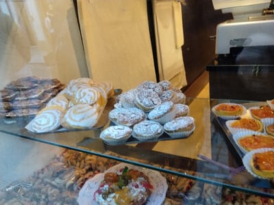 A bakery display case features an assortment of pastries. On the top shelf are stacks of thin, dark-colored pastries, alongside coiled, glazed buns. Small, round pastries dusted with powdered sugar sit next to a tray of miniature tarts filled with orange marmalade. The lower shelf showcases a variety of treats, including a decorated cake with candied fruits, and a multitude of small cookies and baked goods.