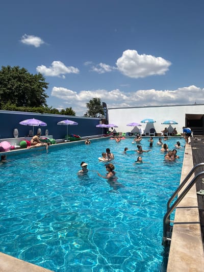 This image depicts a crowded outdoor swimming pool on a sunny day, likely at a resort or recreational facility in Varvara, Bulgaria, given the context. The pool is filled with clear blue water, reflecting the bright sky and scattered white clouds. Numerous people of various ages are enjoying the water, some swimming, others floating, and a few socializing at the poolside.
Along the edge of the pool, several purple umbrellas provide shade. In the background, a blue wall is visible, decorated with what appear to be large, abstract vase-like sculptures and lounge seating. On the right side of the image, a wooden deck area is present with a metal pool ladder providing access to the water. The overall atmosphere suggests a lively summer day with people engaging in recreational activities.