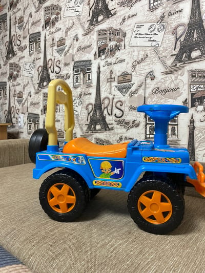 A blue toy jeep with an orange seat and steering wheel is parked on a beige, textured couch. The jeep has a yellow roll bar and orange wheels. A sticker on the side of the jeep depicts a cartoon construction worker. The background is a wallpaper with a pattern of Parisian landmarks, such as the Eiffel Tower and the Arc de Triomphe, along with French text like "Paris" and "Bonjour". There is a white electrical outlet on the wall. The scene appears to be indoors, likely in a home setting, and the location of Бескөл ауылдық округі, Kazakhstan, is not evident from the visual information provided.
