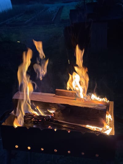 The image depicts a close-up, night-time view of a brazier with burning logs. The fire is actively burning, with bright orange and yellow flames licking upwards. Two logs are visible, stacked horizontally within the brazier. The brazier itself is dark and appears to be made of metal, with decorative cutouts along its top edge. In the background, the scene is dark and indistinct, suggesting an outdoor setting. The overall impression is one of a cozy, outdoor fire. The location is specified as the city of Maksimovo, Russia.