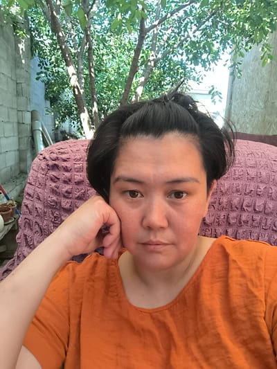 A woman with dark hair pulled back is resting her head on her hand and looking at the camera. She is wearing an orange shirt. She is seated in a textured purple chair outdoors. Behind her, there are trees with green leaves and a brick wall. The lighting suggests it is daytime.