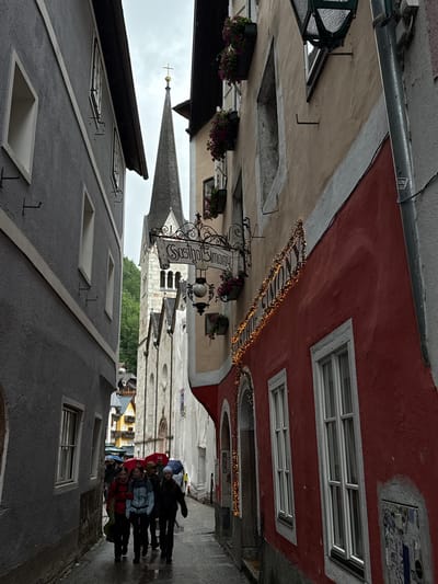The image shows a narrow street in Hallstatt, Austria, on a rainy day. On the left is a grey building with several windows. On the right is a building with a red facade adorned with Christmas lights and a sign that reads "Gasthof Simony". A tall church spire with a golden cross is visible in the background, as well as other buildings with colorful roofs. Several people are walking down the street, holding umbrellas. The ground is wet, indicating recent rainfall.