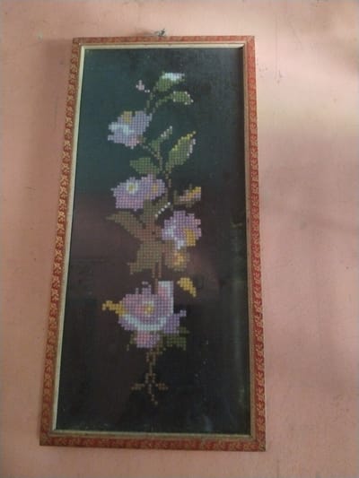 A vertically oriented, framed piece of cross-stitch artwork hangs on a light peach-colored wall. The artwork features a floral design, depicting a vine or stem with several stylized flowers and leaves against a dark, possibly black, background. The flowers are rendered in shades of light purple, lavender, and white, with small yellow centers. The leaves are predominantly green with some yellowish-green accents. The cross-stitch technique is evident, showing the individual stitched "pixels" that form the image.

The frame is rectangular, with a reddish-brown base color and a golden, intricate patterned border that appears to repeat a small floral or geometric motif. A small string or wire used for hanging is visible at the top center of the frame, attached to a hook or nail on the wall. The wall itself shows some faint discolorations and marks, suggesting an aged or lived-in environment. There is no discernible activity taking place, as it is a static display of an indoor decoration. No text is visible in the image. The scene is likely indoors in Teupinbayu, Indonesia.