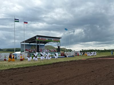 The image depicts an outdoor festival or celebration at the city of Nurkeevo, Russia, under a cloudy sky. A stage is set up on the right side of the frame, with banners that read "Нуркеево 2026" and other text in Cyrillic. Several flags are visible, including the Russian flag and another flag with horizontal stripes of blue, white, and green.
In front of the stage, a group of performers, predominantly women and girls, are engaged in a dance. They are dressed in traditional colorful attire, with some wearing white dresses and veils, while others are in vibrant yellow and red garments. They are arranged in lines and circles, performing synchronized movements.
To the left of the stage, more performers in traditional clothing stand, some holding circular props with patterns. Spectators are visible in the background, near the stage and along the perimeter of the performance area.
The environment consists of a grassy field with a strip of freshly tilled brown earth in the foreground, suggesting a rural or park-like setting. Trees and rolling hills form the backdrop behind the stage, with a few scattered structures visible in the distance. The overall atmosphere appears festive and communal.