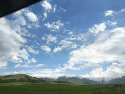 This media file is a landscape photograph taken from a moving vehicle, likely a car, as indicated by the dark frame in the upper left corner, which appears to be part of the vehicle's interior. The scene depicts a vast, rolling green landscape under a bright blue sky dotted with numerous white, fluffy cumulus clouds.
The foreground is dominated by expansive fields of vibrant green grass, suggesting a rural or agricultural setting. In the mid-ground, rolling hills rise, some covered with dense evergreen trees forming distinct patterns and shapes. The farthest hills in the background are shrouded in mist or low-lying clouds, creating a hazy, atmospheric effect and obscuring their peaks. Two utility poles with wires are visible in the field, and to the far right, a small, unfinished concrete structure with two openings, possibly a shed or an animal enclosure, is present. A faint outline of what could be a cow is visible near the structure.
The time of day appears to be daytime, with bright sunlight casting shadows and illuminating the landscape. The weather is fair, with a mix of sun and clouds, typical for a pleasant day. The location is identified as Alagyaz, Armenia, which is known for its mountainous terrain and agricultural plains. The overall mood conveyed by the image is one of natural beauty, tranquility, and openness, evoking a sense of journey and exploration. There is no visible text in the image.