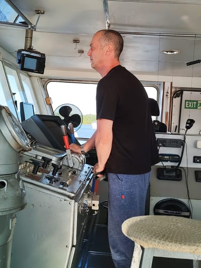 The image shows a man standing in the pilot house of a vessel, appearing to be at the helm. He is wearing a black t-shirt and blue sweatpants, and is looking to his right, out a window. His hands are on a control lever system, suggesting he is operating the vessel.
The pilot house is equipped with various instruments and controls, including a large compass to the left, a series of levers and dials in the center, a fan behind the controls, and communication equipment on the right wall, including a microphone and a radio. A screen is mounted on the ceiling, and an "EXIT" sign is visible on the right wall. The interior appears functional and well-used, consistent with a working vessel.
Through the windows, a body of water and a tree-lined shore are visible, indicating the vessel is likely on a river or lake. The lighting suggests it is daytime, with bright natural light illuminating the scene. The overall atmosphere is one of a person diligently performing their duties. The setting is identified as Darghun, Russia.