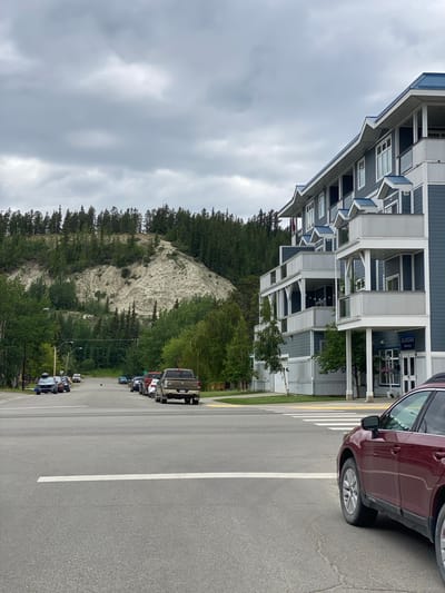 The image shows a street scene in Whitehorse, Canada, with a focus on a multi-story building on the right and a tree-covered hillside in the background. The building, with its dark blue siding and white trim, appears to be an apartment complex or condominium, featuring multiple balconies with white railings and blue roofs. A sign above the entrance reads "AURORA Wellness Group."
The street in the foreground is paved with asphalt and marked with white lines, indicating a crosswalk. A dark red SUV is parked in the foreground, partially obscuring the view of the crosswalk. On the left side of the street, several other vehicles are parked along the curb, including a silver SUV, a dark blue sedan, and a pickup truck.
The background features a steep, light-colored hillside covered with a dense forest of evergreen trees. The sky is overcast, with heavy gray clouds dominating the upper portion of the image. The overall impression is that of a residential area in a northern climate, possibly during the summer or shoulder season, given the foliage and the overcast sky. There are no people clearly visible in the image, and the focus is on the architecture, vehicles, and natural landscape.