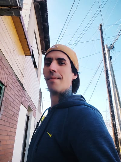 A low-angle, rotated portrait shows a person wearing a dark blue hooded sweatshirt and a tan baseball cap. The person is looking towards the camera with a neutral expression. Tattoos are visible on their neck. The background consists of the side of a building with exposed brickwork and yellow siding, and a bright blue sky crisscrossed with utility wires. The lighting suggests it is daytime, likely sunny, with no indication of specific weather conditions. The scene appears to be outdoors, possibly in an urban or semi-urban environment, given the building and the dense network of wires. No visible text is present in the image.